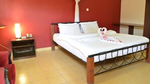Una cama bien hecha con sábanas blancas y un arreglo decorativo de almohadas con forma de cisne. Se ve una mesita de noche con una lámpara y un teléfono. La habitación tiene una pared roja y un suelo de baldosas.
