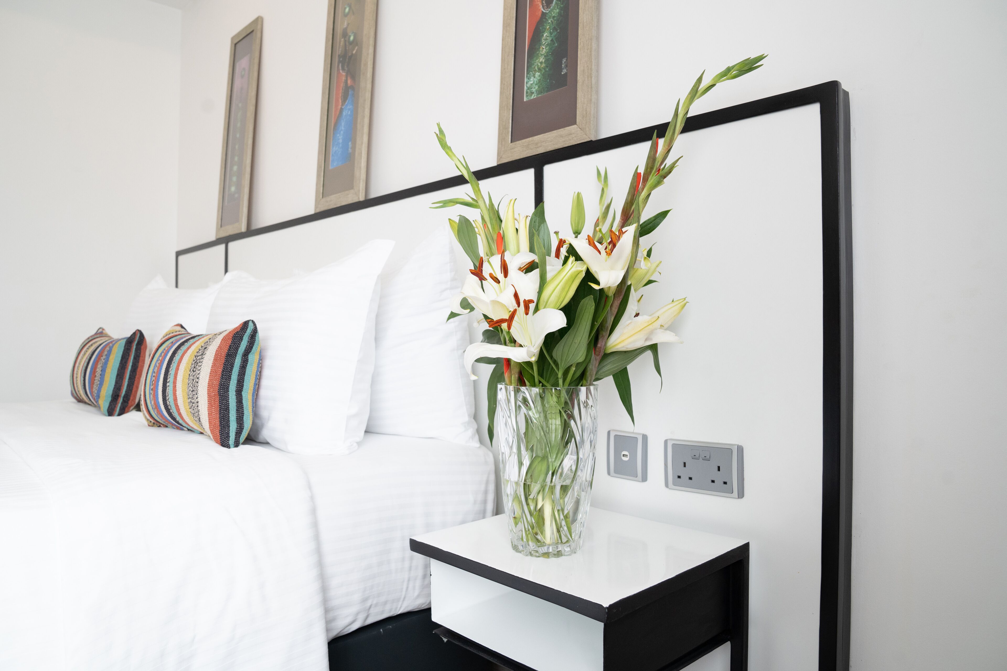 A neatly made bed with white bedding and colorful pillows, a clear vase with white flowers on a bedside table, and a wall with framed artwork.