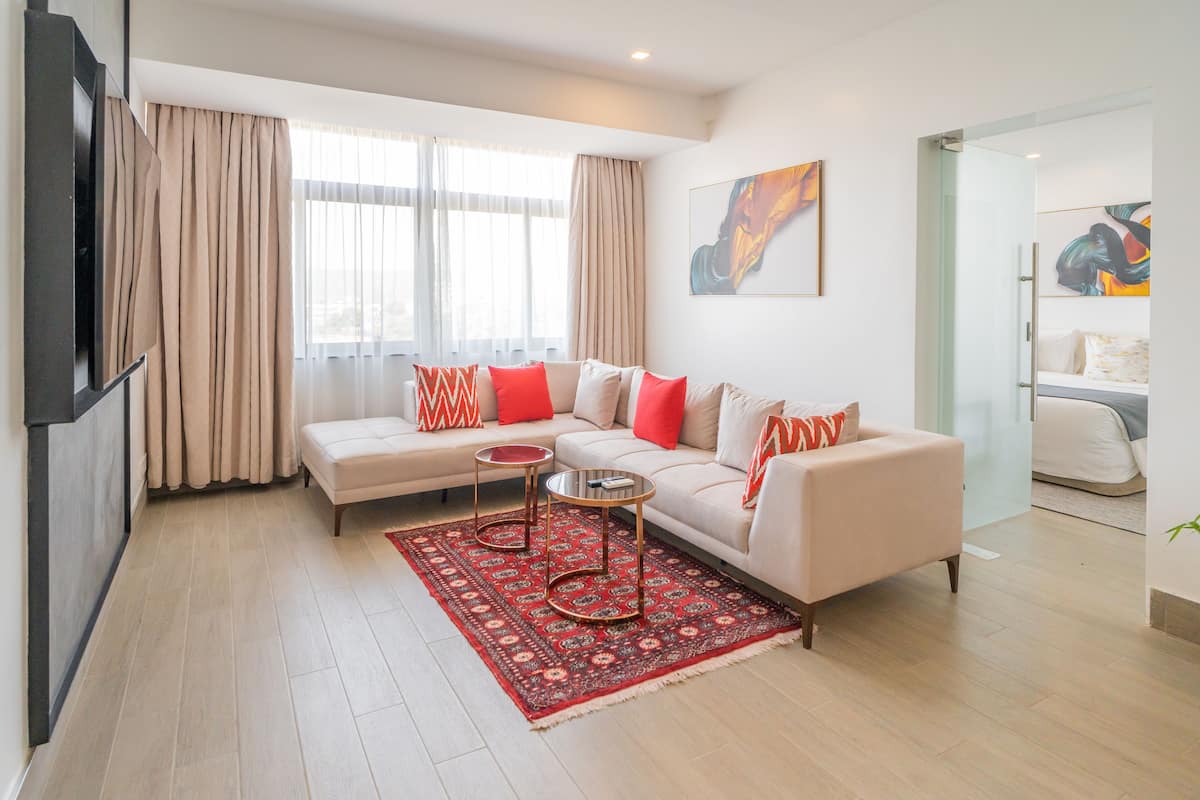Una sala de estar moderna con un sofá beige, cojines rojo y blanco, una mesa de centro de vidrio y una alfombra roja estampada.