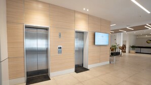 Un área de ascensores moderna con dos ascensores, una pantalla digital y un mostrador de recepción en un vestíbulo limpio y bien iluminado.