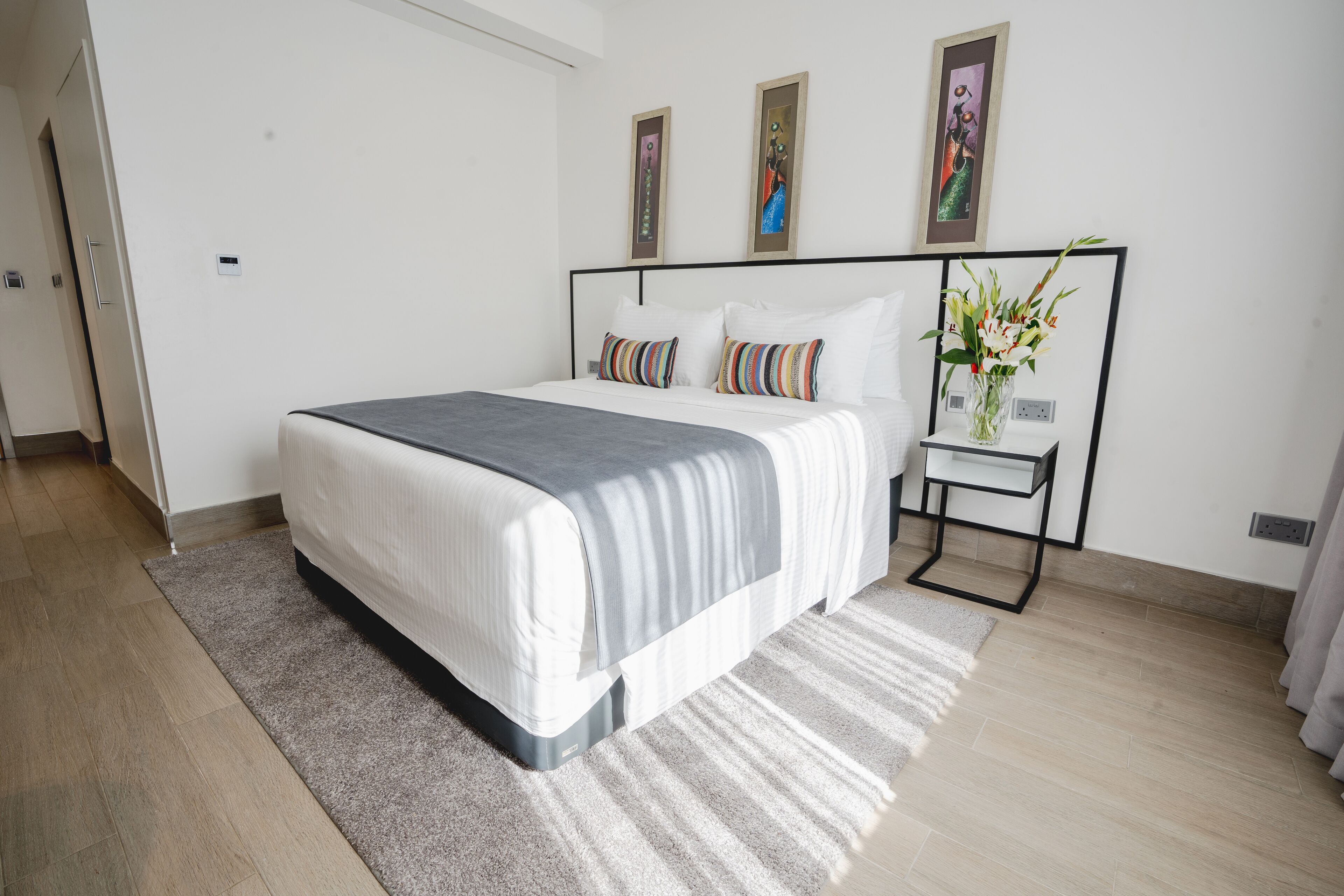 A modern bedroom with a large bed, a bedside table with a vase of flowers, and two framed artworks on the wall.