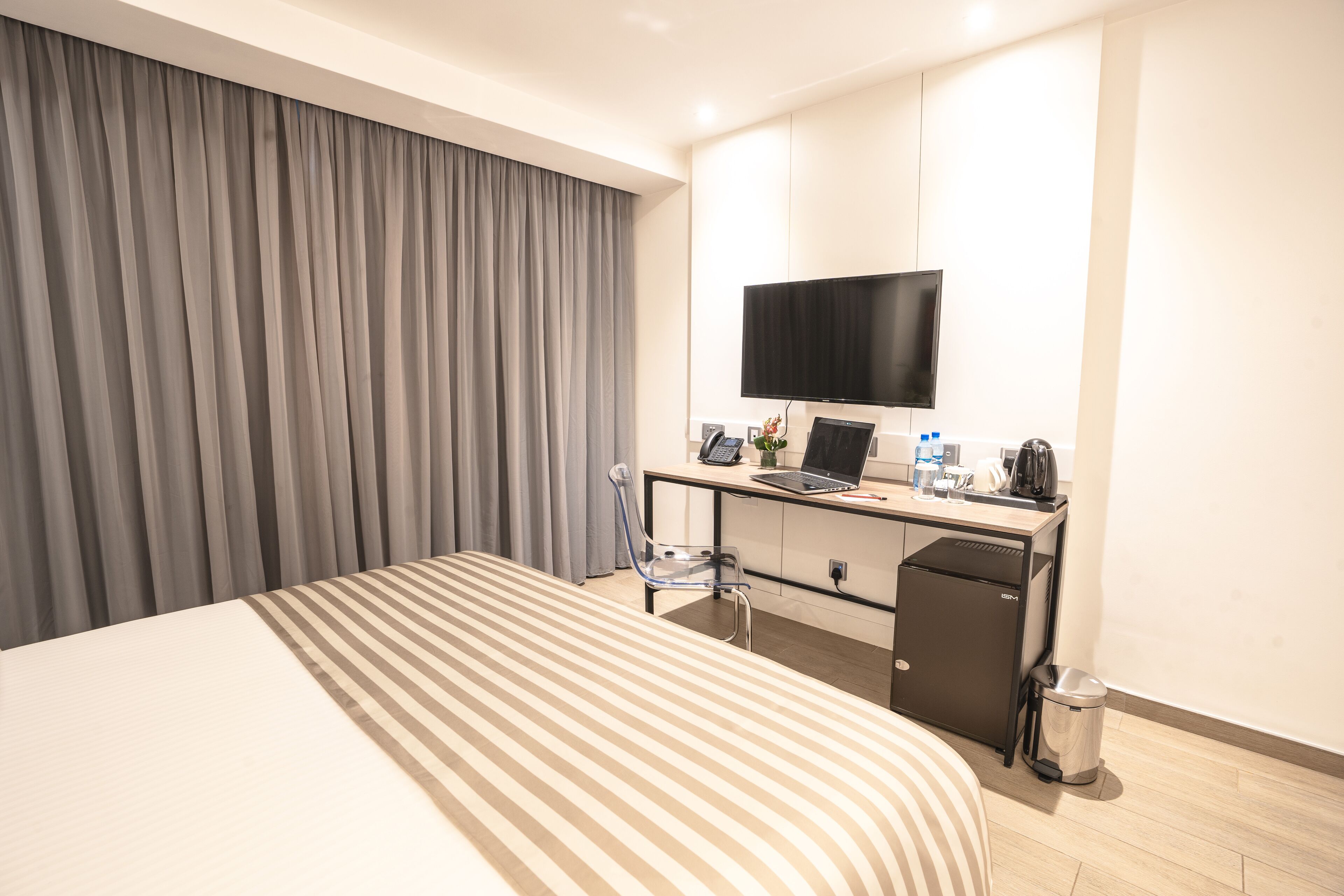 A hotel room with a bed, a desk with a laptop, a television, and a trash can.