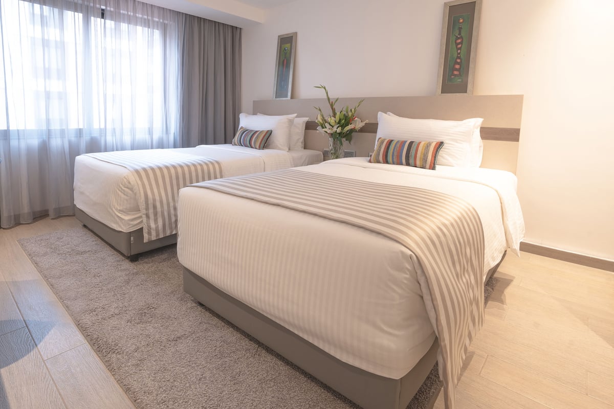 Habitación de hotel con dos camas, un ventanal con cortinas, un jarrón con flores y un cuadro en la pared.