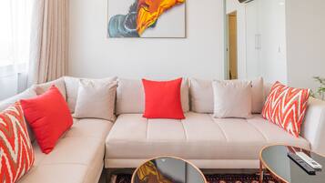 Een moderne woonkamer met een beige bank, rode en gemêleerde kussens, een glazen salontafel, en een kleurrijk abstract schilderij aan de muur.