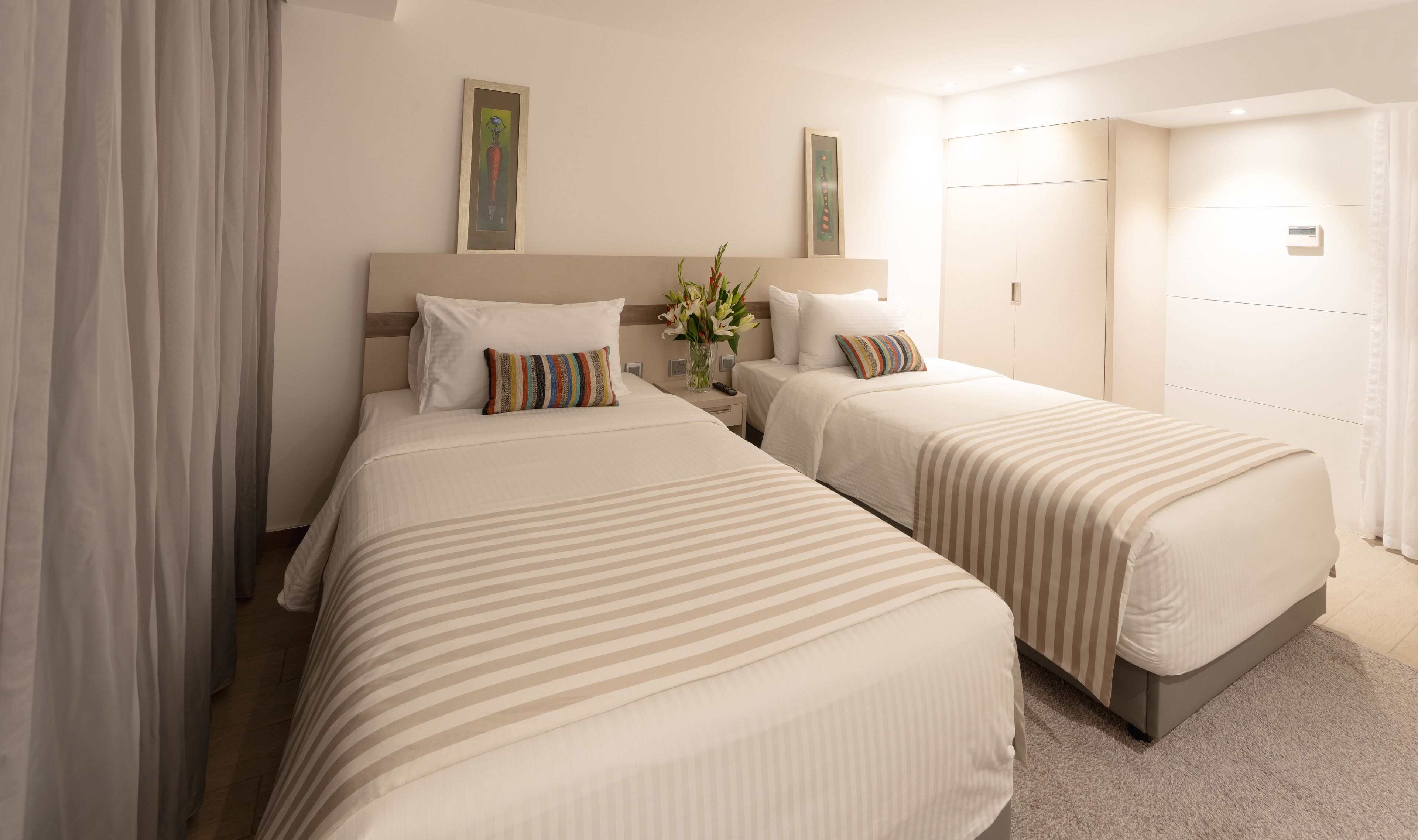 A hotel room with two beds, a nightstand, a vase with flowers, and a painting on the wall.