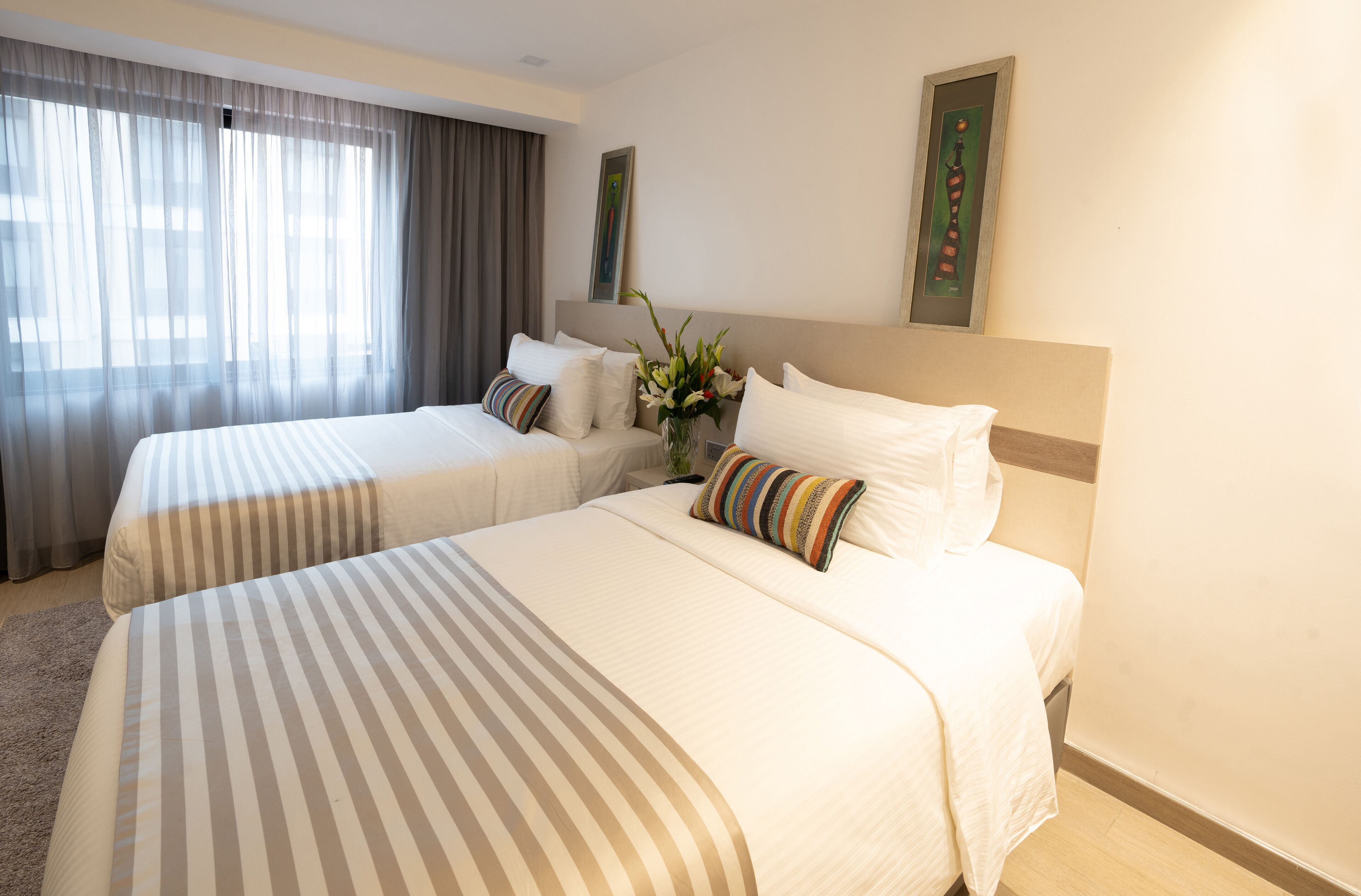 Habitación de hotel con dos camas, un ventanal con cortinas, un jarrón con flores y un cuadro en la pared.