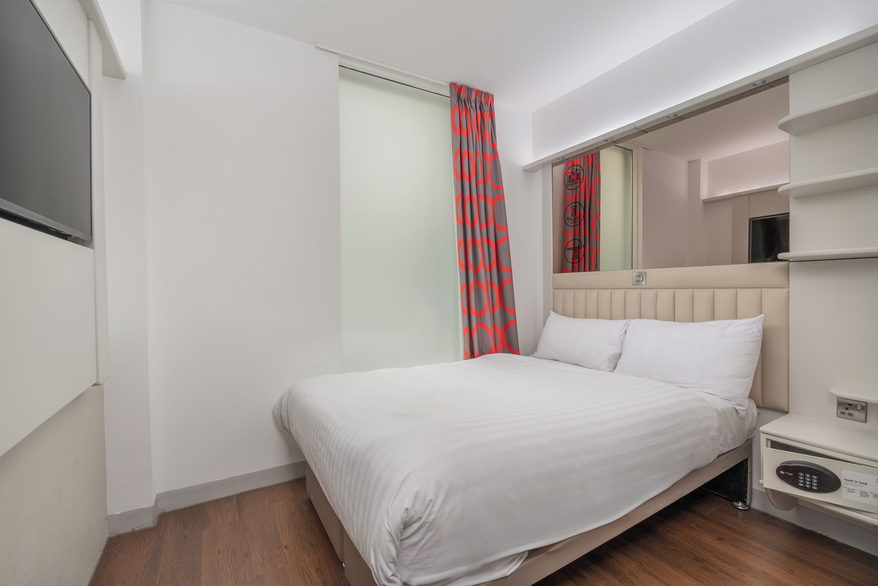 A hotel room with a bed, a bedside table with a control panel, a mirror above the bed, and a sliding door with a red and gray curtain.