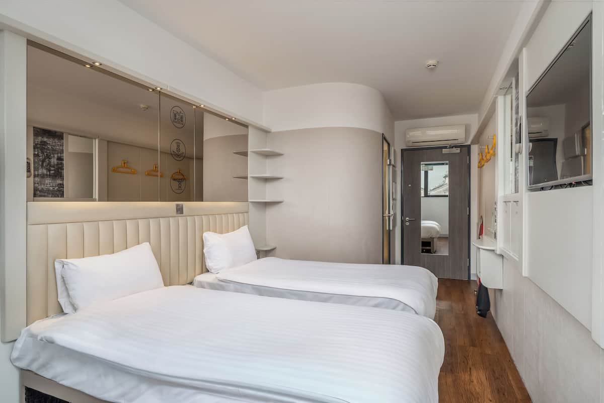 A hotel room with two beds, a large mirror, a wardrobe, and a view of the city.