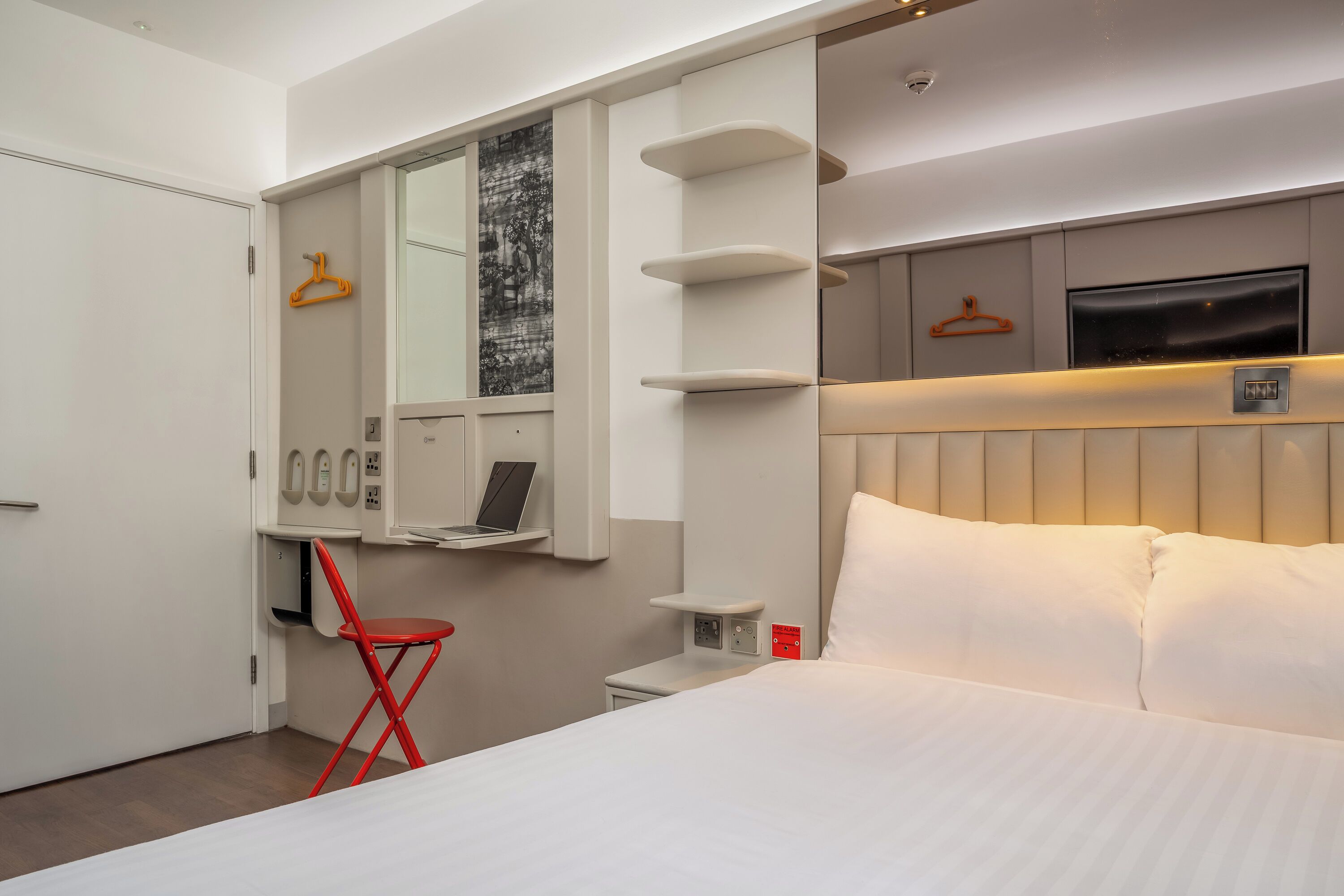 Une chambre d’hôtel avec un lit, un bureau avec un ordinateur portable, une chaise rouge, une télévision et une étagère fixée au mur.