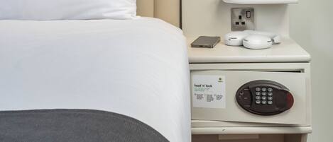 Una habitación de hotel con cama, mesita de noche con teléfono, cafetera y estante con libros y un ordenador portátil.