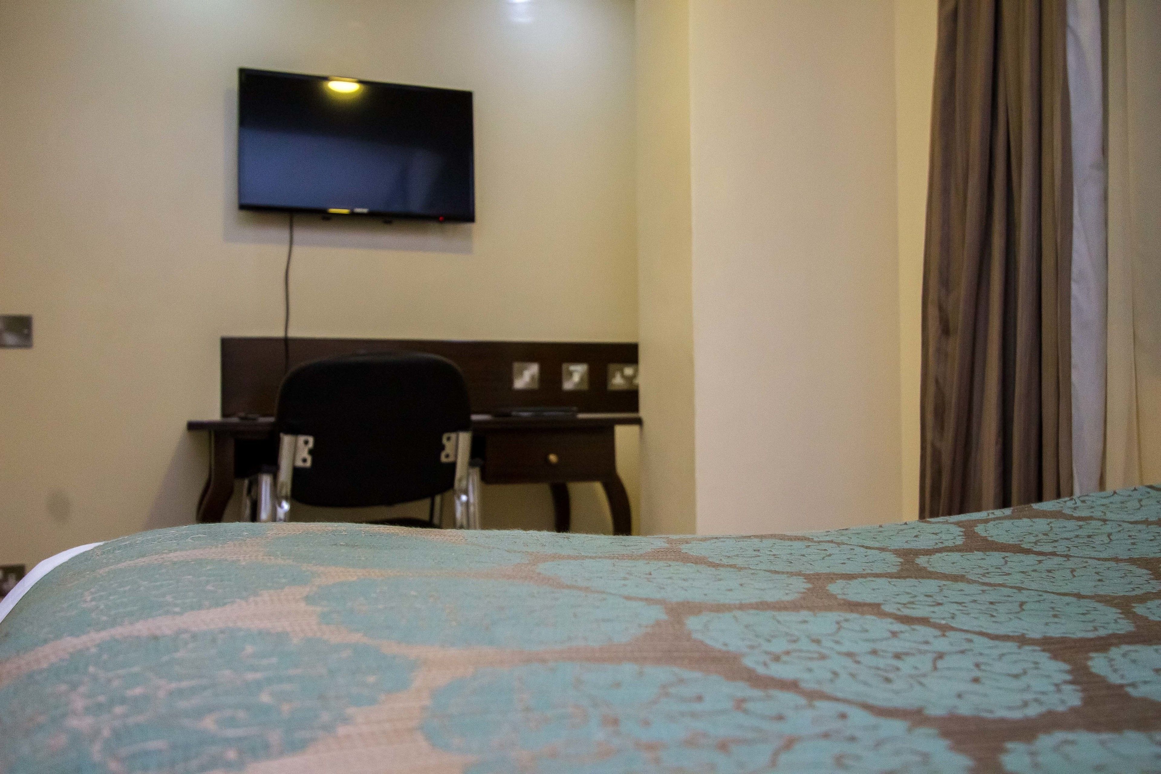 A hotel room with a bed, a desk, a chair, a television mounted on the wall, and a curtain on the window.