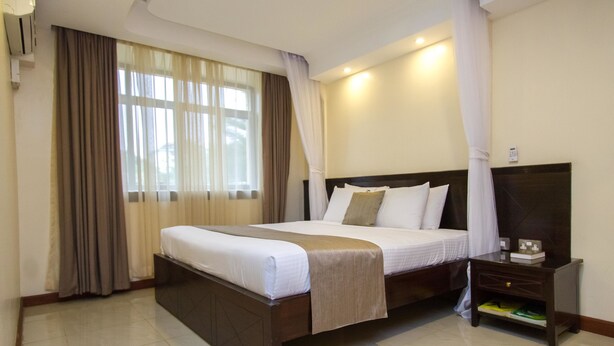 A hotel room with a large bed, a nightstand, a TV, and a window with curtains. - Pinecone Hotel (Kisumu)