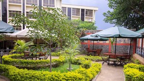 An outdoor courtyard with tables and chairs under umbrellas, surrounded by well-maintained hedges and a central tree. - Pinecone Hotel (Kisumu)