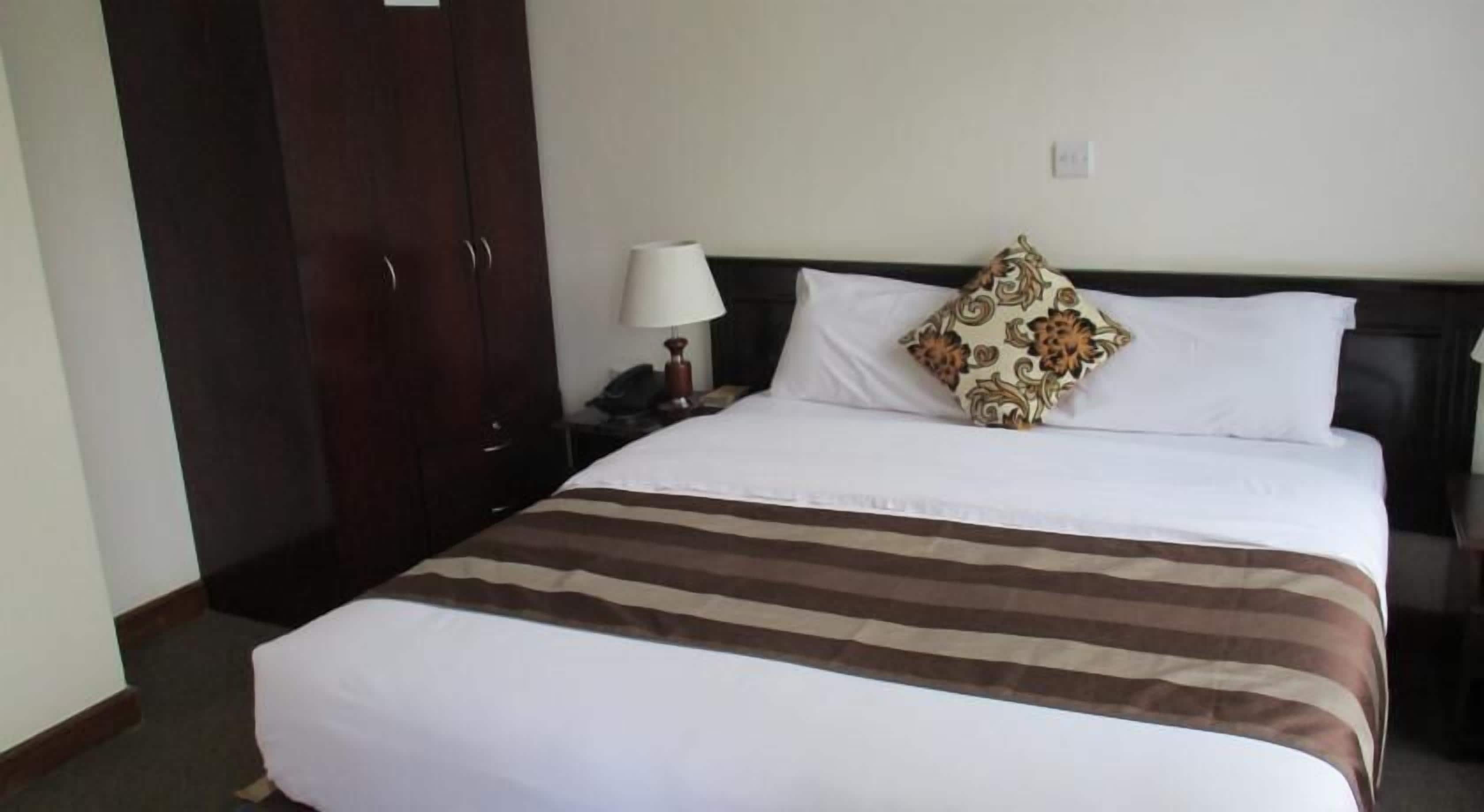Una habitación de hotel con una cama grande, una mesita de noche con lámpara, un armario y un cojín decorativo.