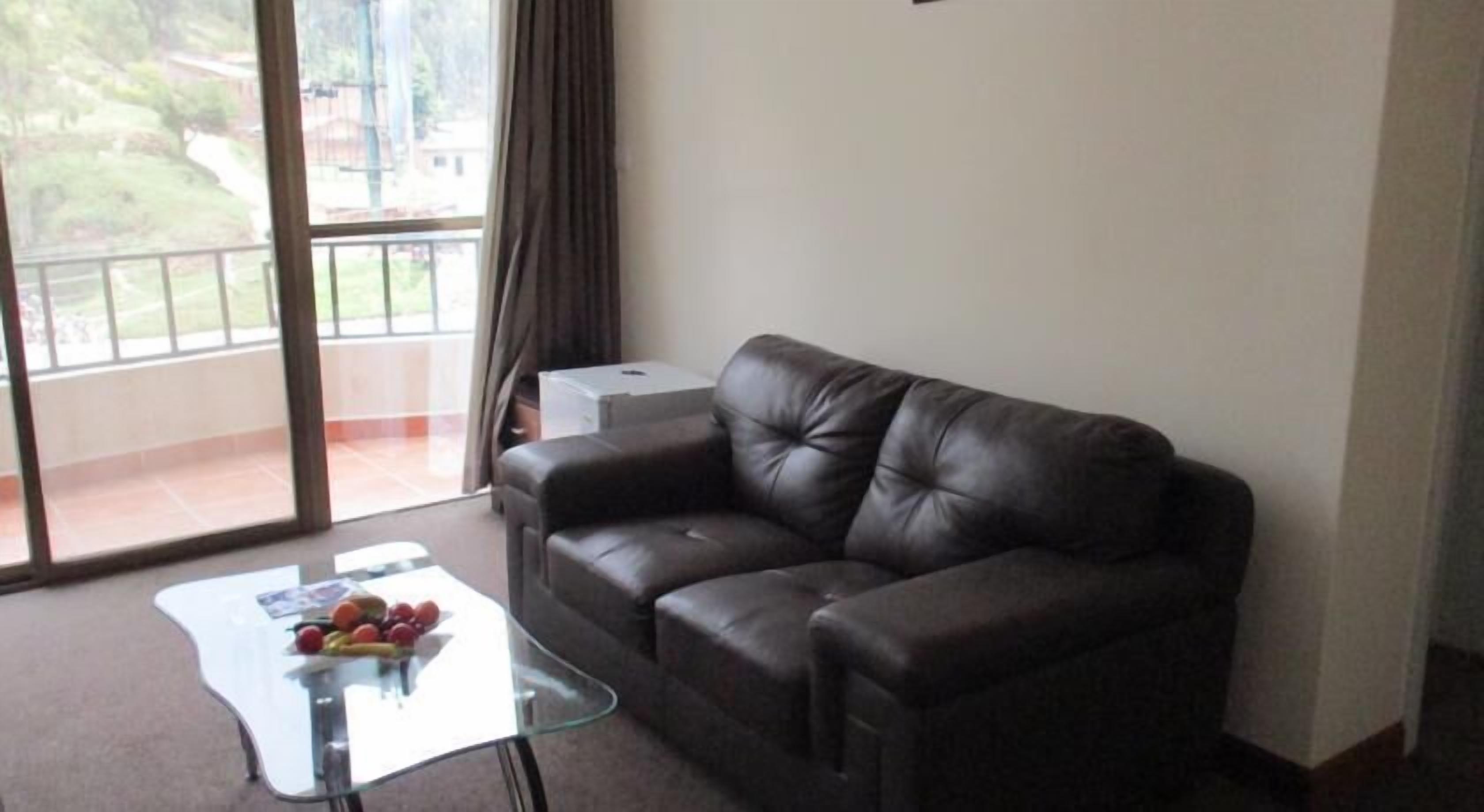 A living room with a leather sofa, a glass coffee table with fruits, and a view through large windows.