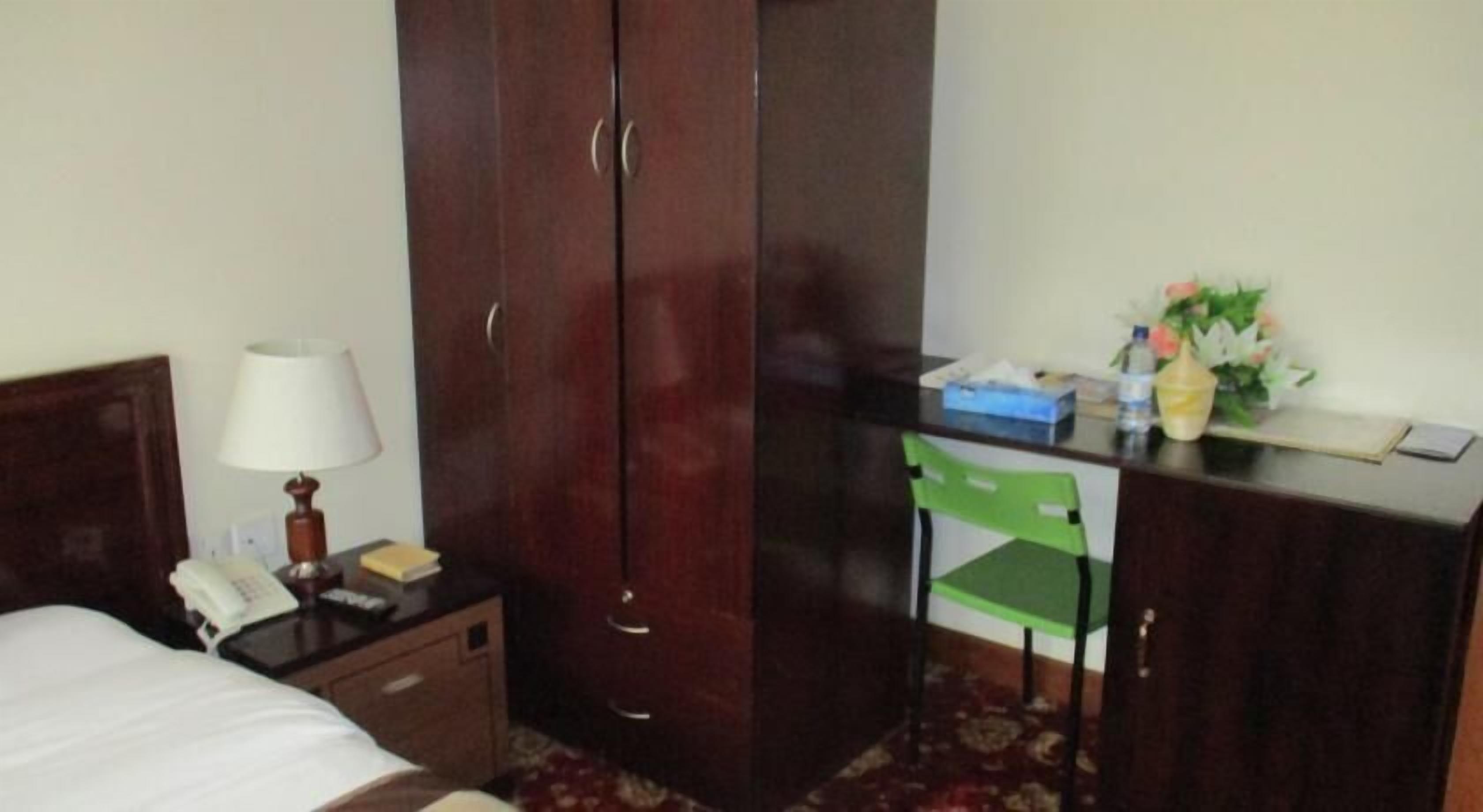 A hotel room with a bed, a desk with a lamp, a green chair, and a wardrobe.
