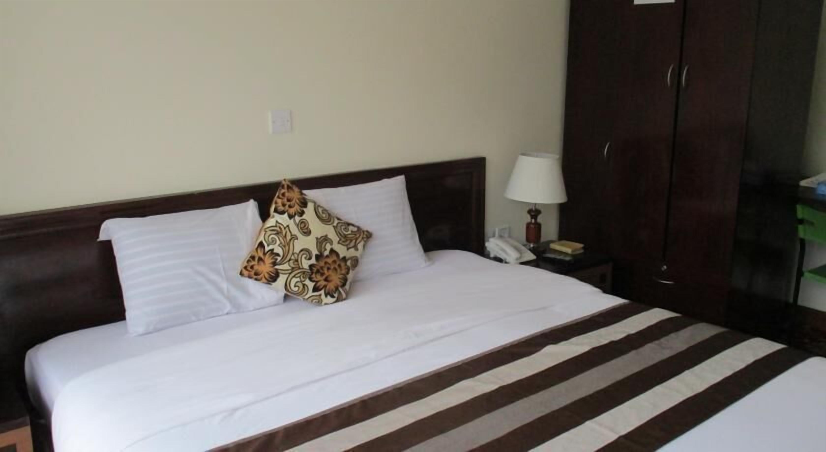 Una cama bien hecha con ropa de cama blanca y a rayas, una almohada estampada, una lámpara de noche y un teléfono sobre una mesita de madera.