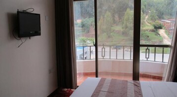 Una habitación con cama, televisor montado en la pared y balcón con vistas a un paisaje verde y una carretera.