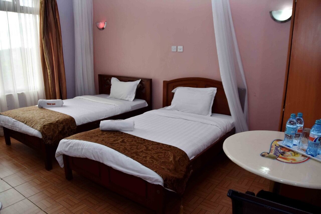 A hotel room with two single beds, a small round table with bottled water, and a window with sheer curtains.