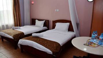 A hotel room with two single beds, a small round table with bottled water, and a window with sheer curtains.