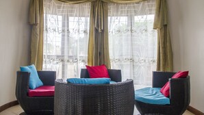 A corner seating area with wicker chairs and cushions, a window with sheer curtains, and a decorative metal screen. - Spannet Suites (Mbarara)