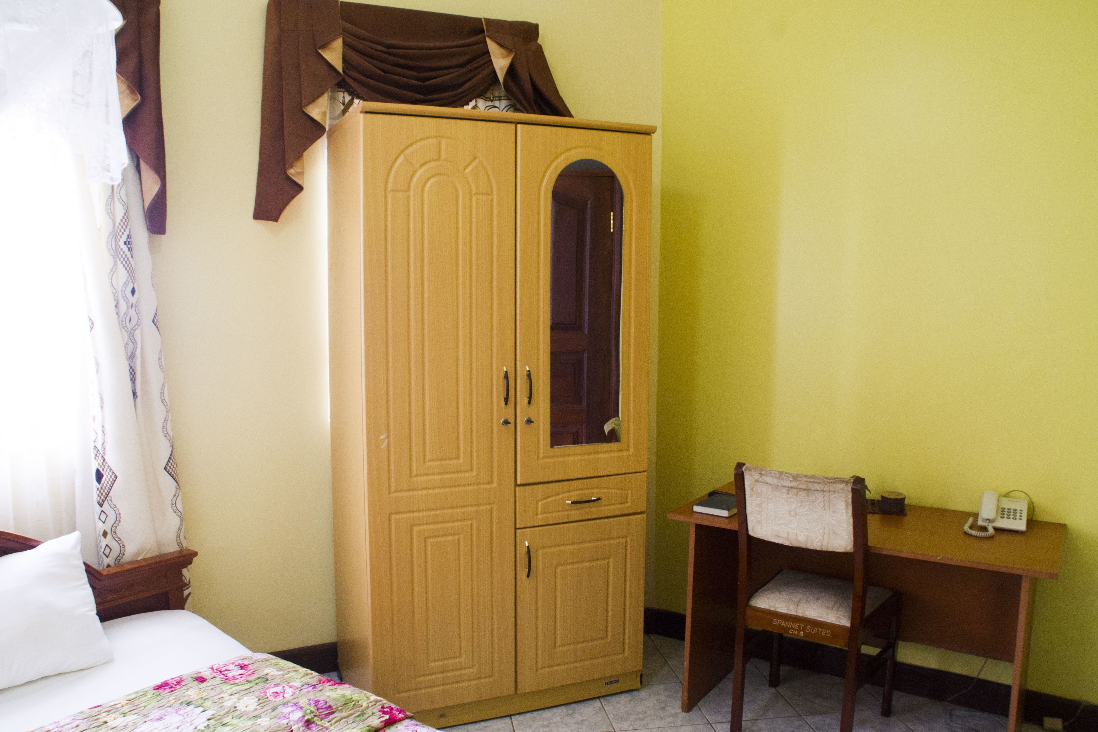 A bedroom with a bed, a wooden wardrobe, a desk with a chair, and a telephone.