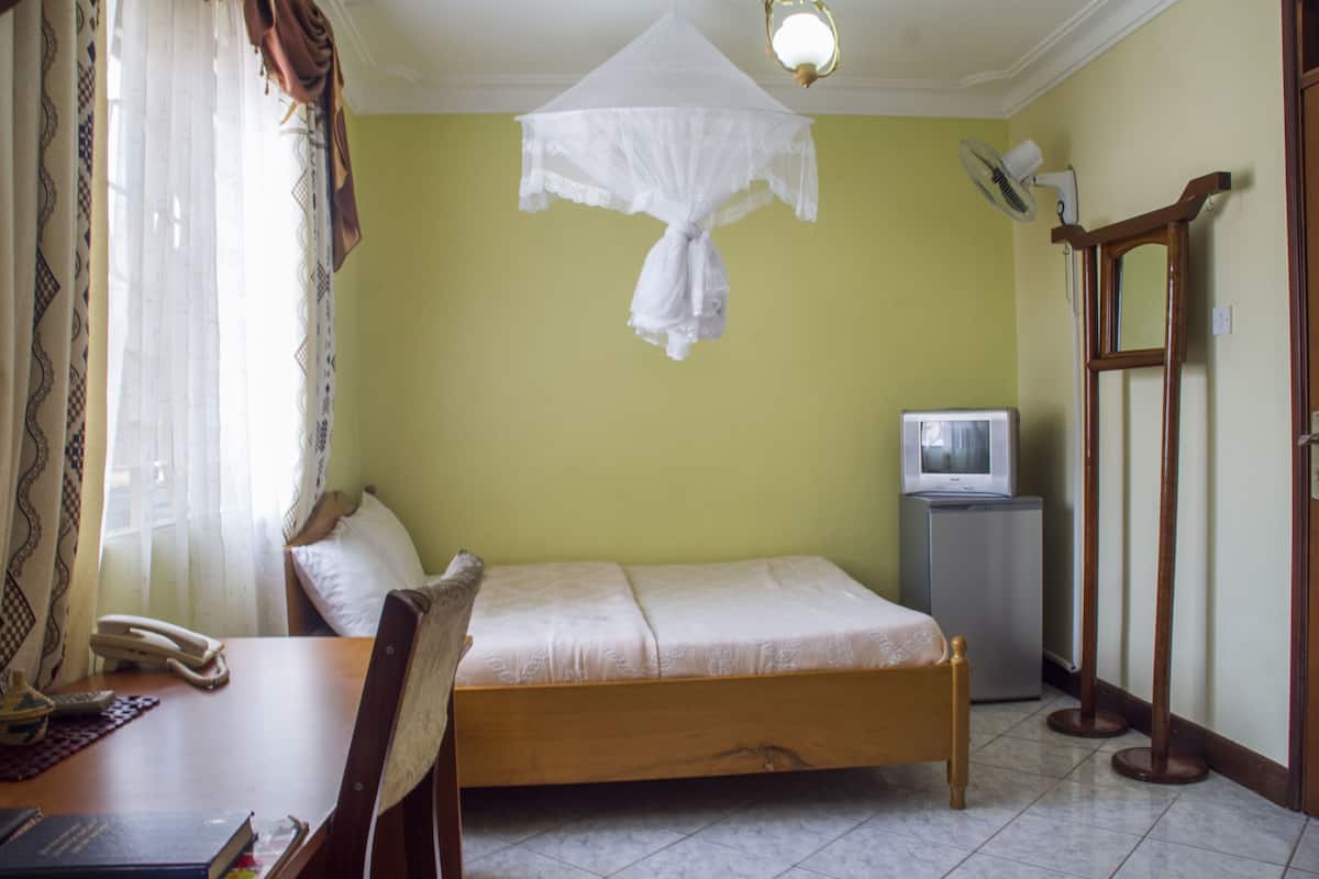 A room with a bed, a desk with a telephone, a television on a stand, and a standing fan.