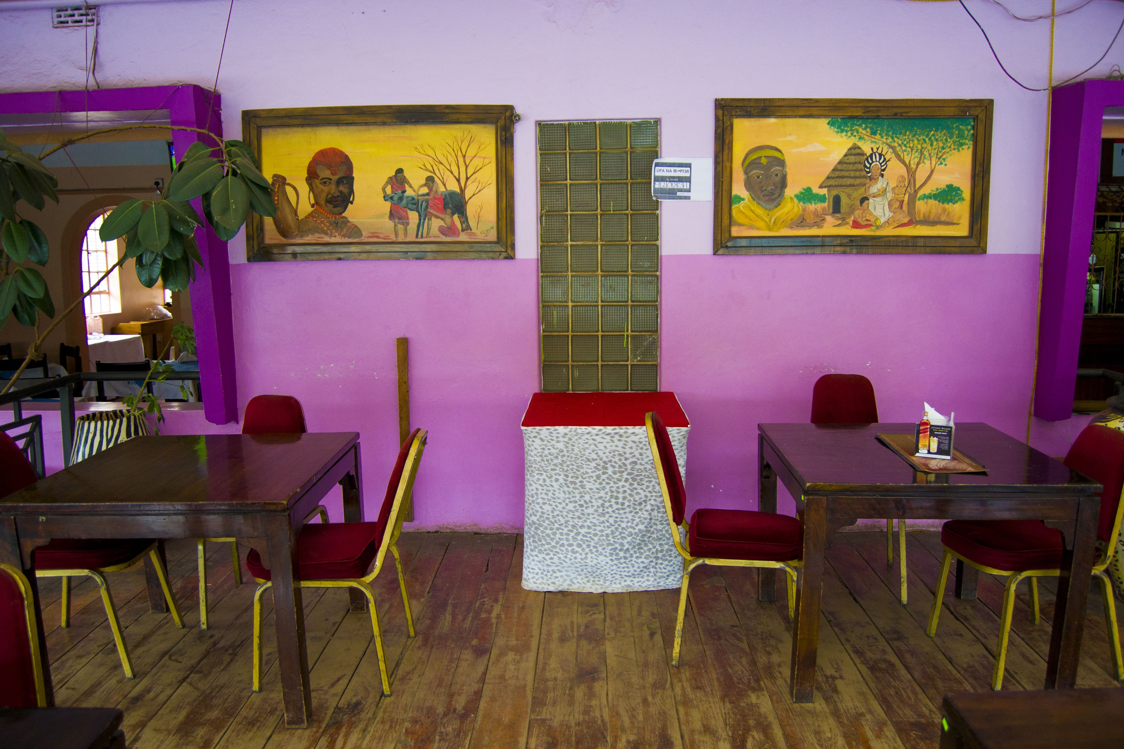 A dining area with wooden tables and red chairs, a small table with a white tablecloth, and two paintings on the wall.