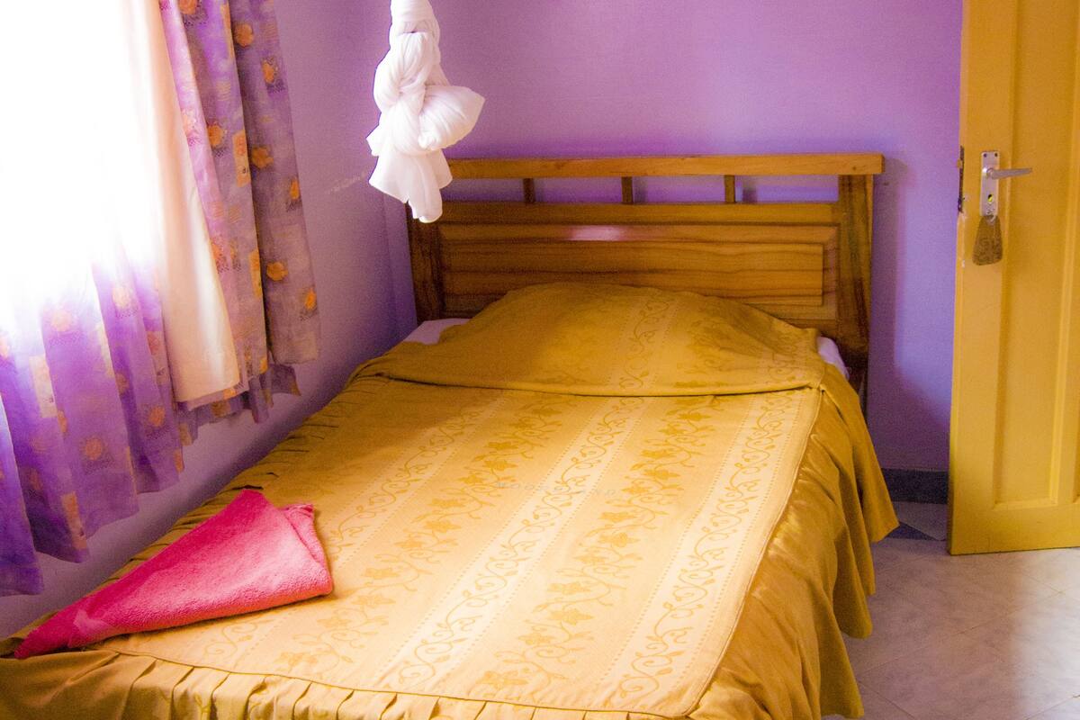 Un lit simple avec une couvre-lit jaune et un ciel de lit blanc, une tête de lit en bois et un mur violet.