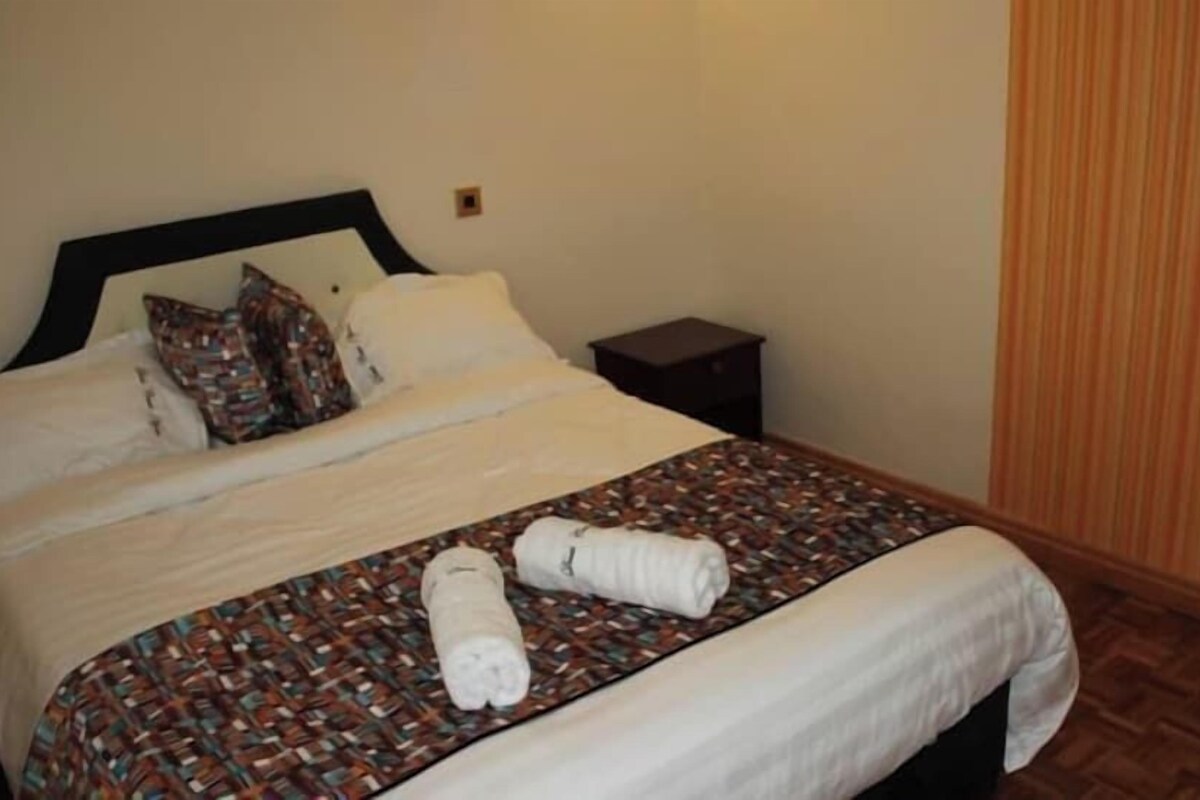 A neatly made bed with a patterned bedspread and pillows, a small bedside table, and a striped curtain.