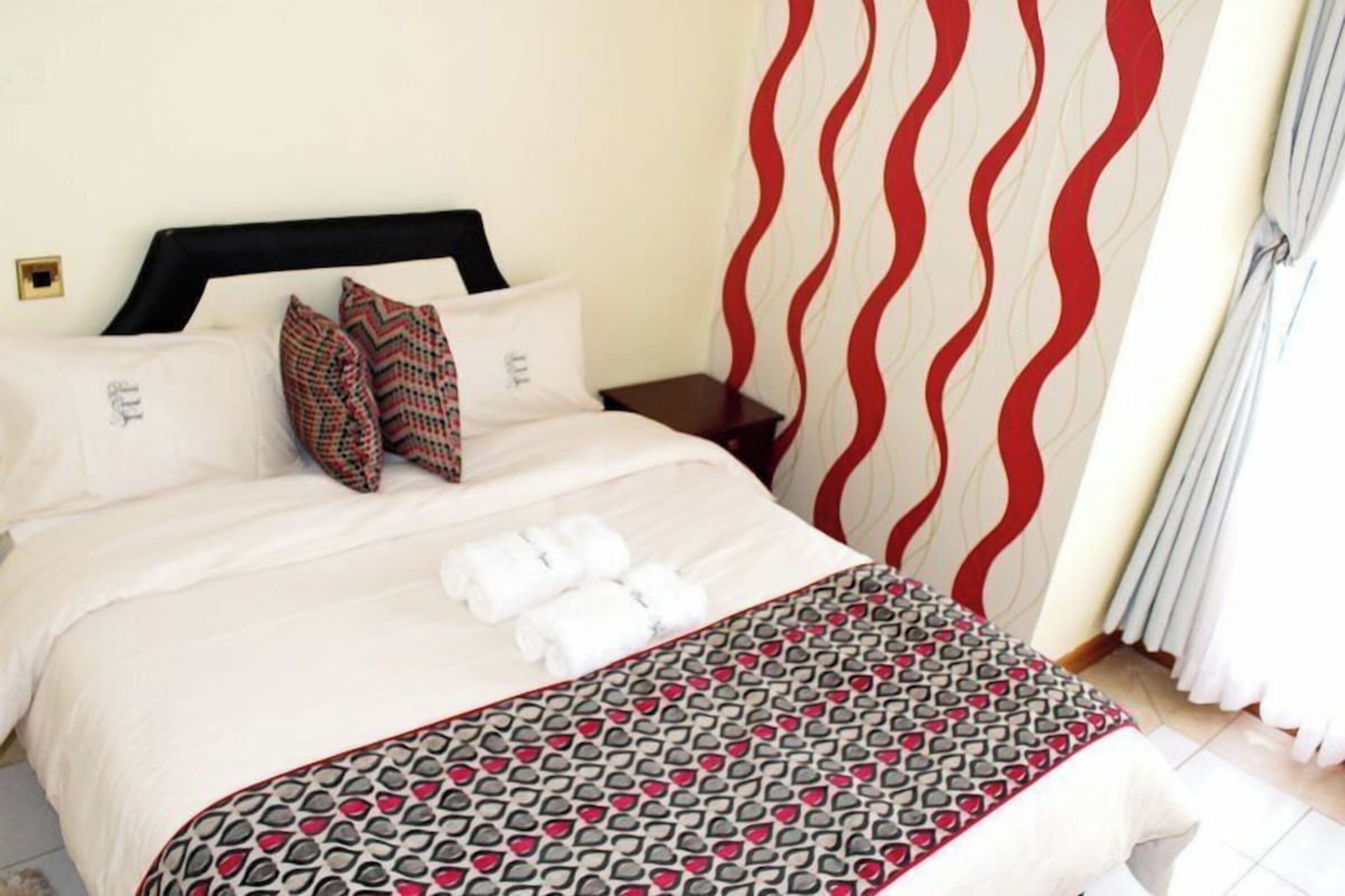 A neatly made bed with white linens and a patterned throw blanket, a nightstand with a lamp, and a red and white wavy curtain.