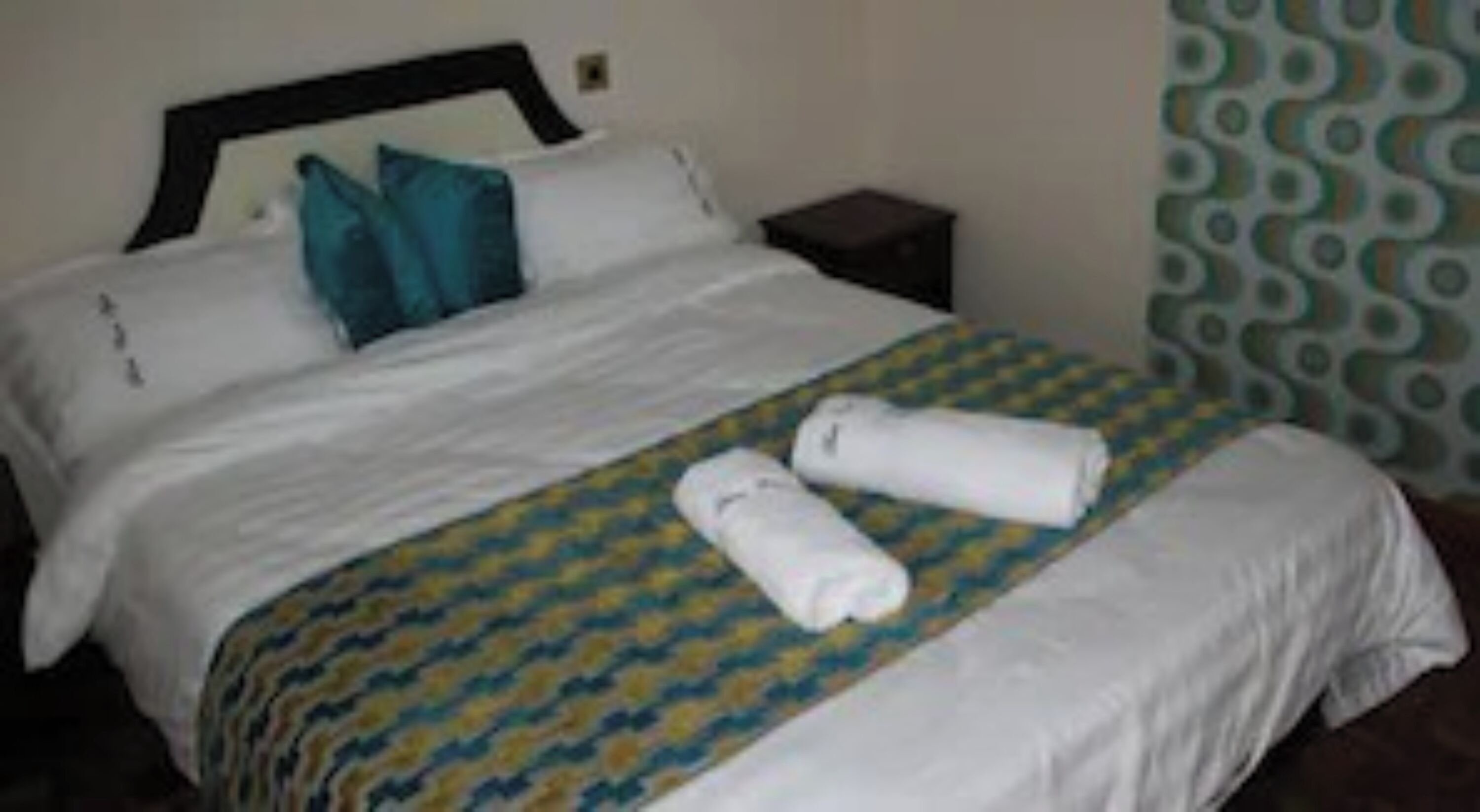 A neatly made bed with a patterned bedspread, two rolled towels, and a nightstand with a lamp.