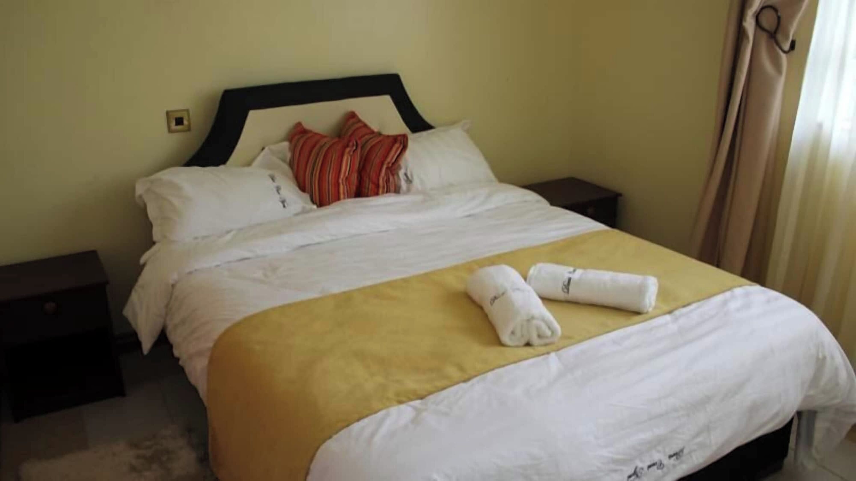 Una cama bien hecha con ropa de cama blanca y amarilla, dos almohadas y dos toallas enrolladas sobre la cama.
