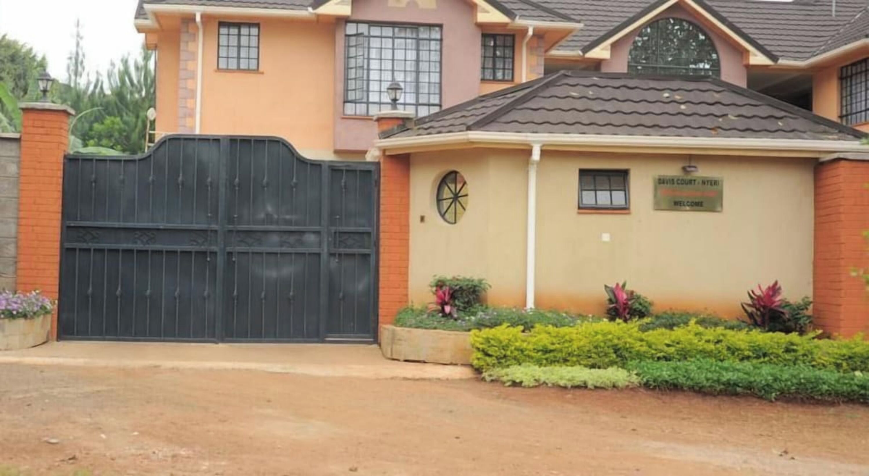 A two-story house with a black gate, a sign reading "Davis Court - Nyeru Welcome", and a small garden with various plants.