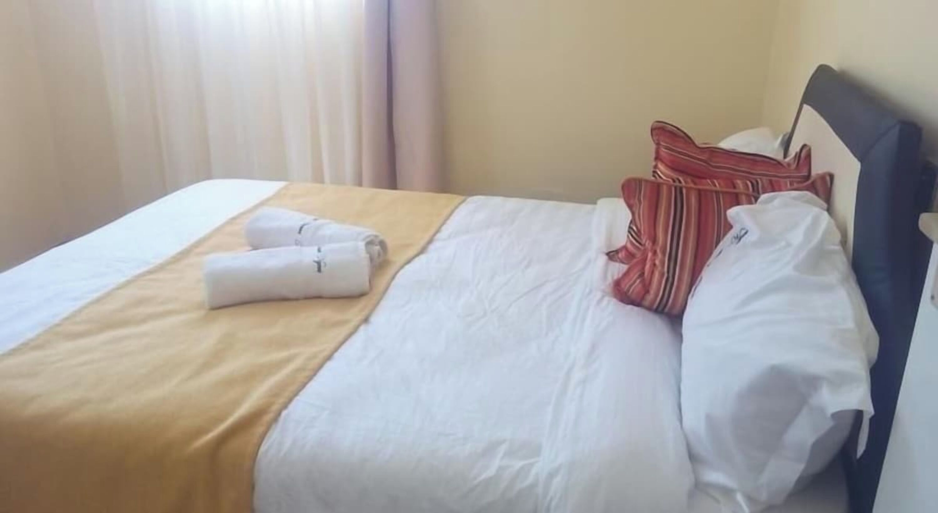 A neatly made bed with white linens, a beige bedspread, and two rolled towels. There are striped pillows and a plain white pillow on the bed.