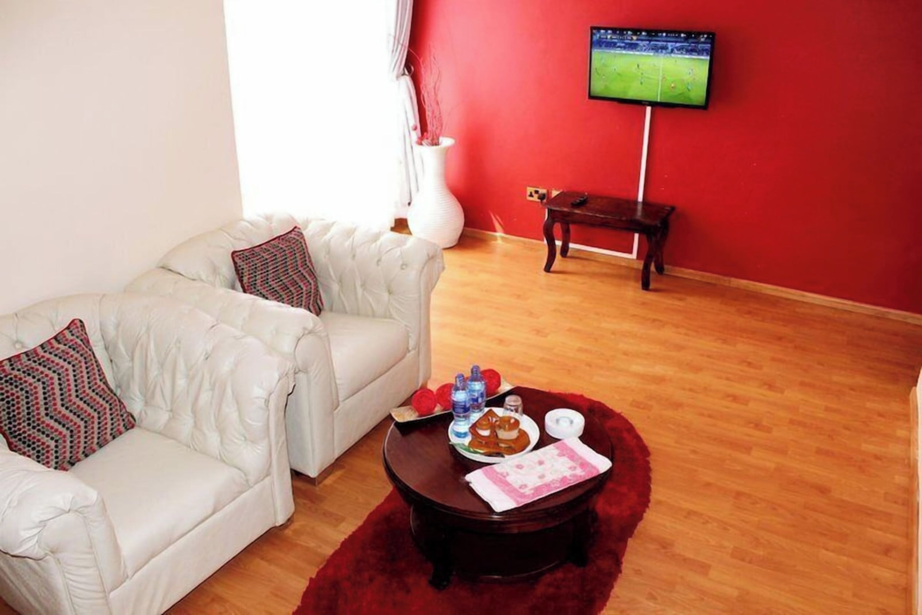 Uma sala de estar com um sofá branco, uma mesa de centro com petiscos, uma televisão na parede e piso de madeira.