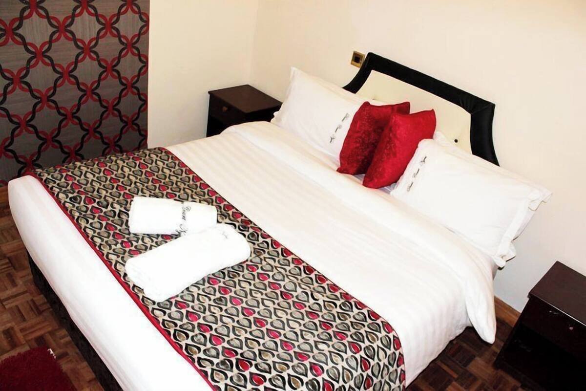 A neatly made bed with a patterned bedspread, white pillows, and a red throw. There are two bedside tables, one with a lamp and the other without.
