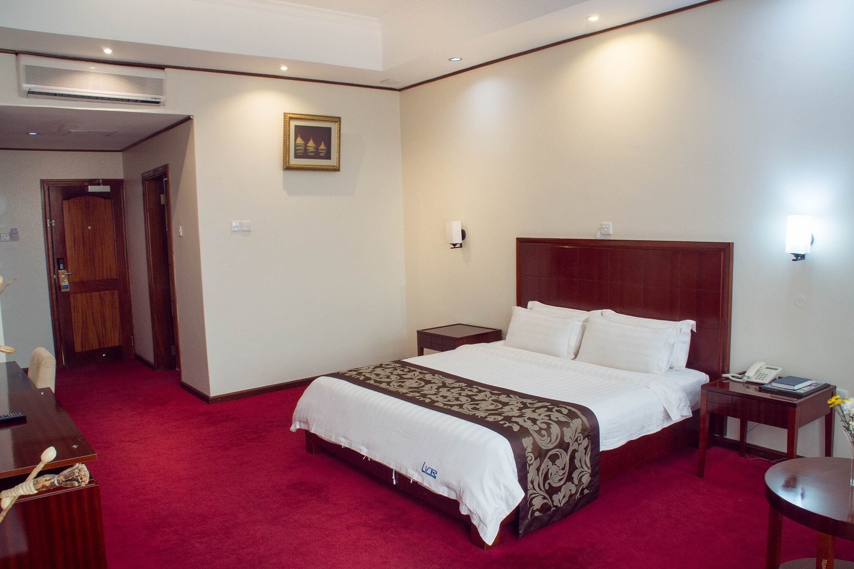 Habitación de hotel con una cama grande, un escritorio y una silla. Se observa una puerta que conduce a otra habitación y un cuadro en la pared.