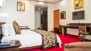 A hotel room with a bed, bedside table, desk, chair, TV, and a door leading to another room. - Lake View Resort Hotel (Mbarara)