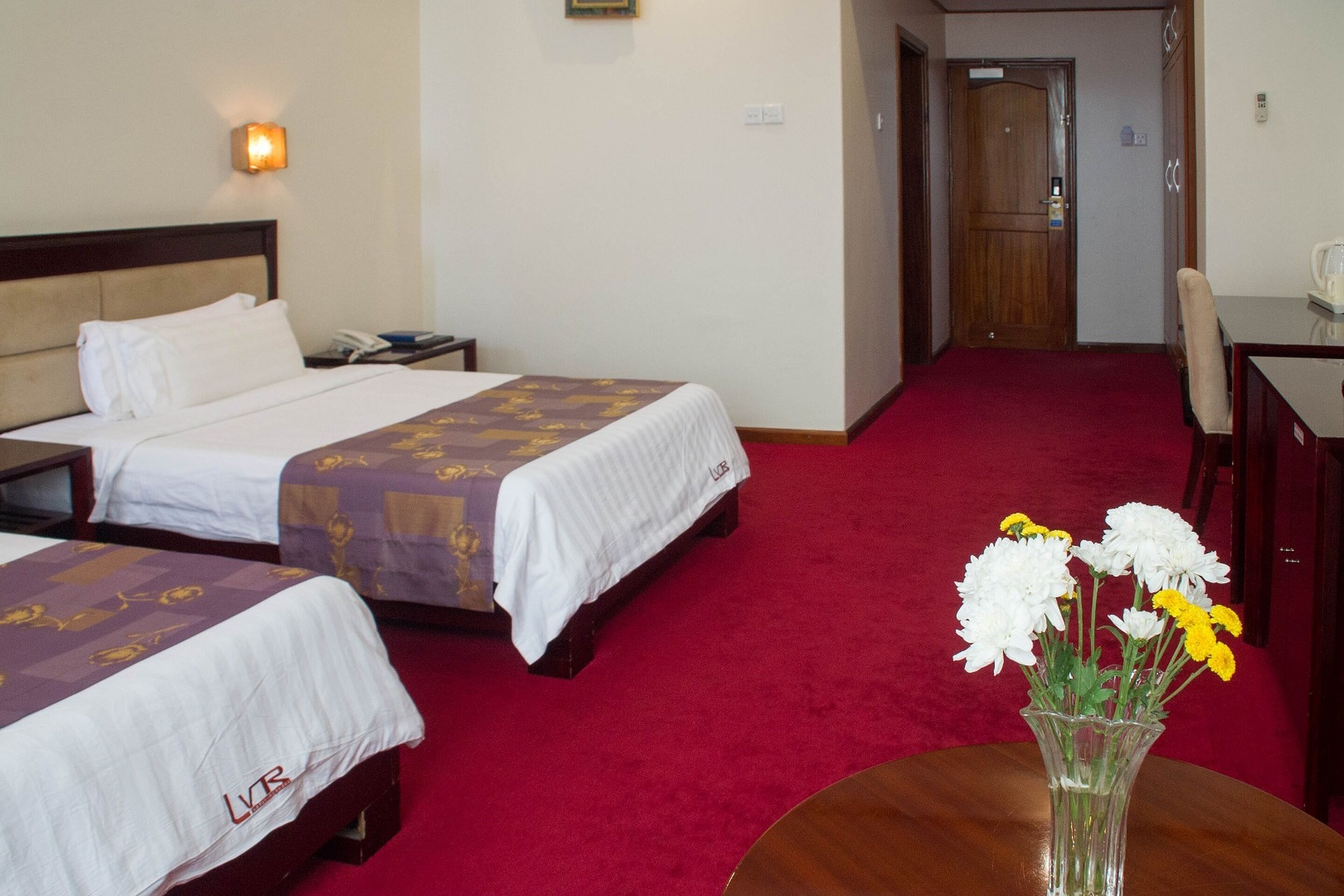 Ett hotellrum med två sängar, ett skrivbord och en stol. På skrivbordet står en vas med blommor.
