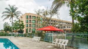 A swimming pool area with lounge chairs and a red umbrella, surrounded by a fence and palm trees. - Lake View Resort Hotel (Mbarara)