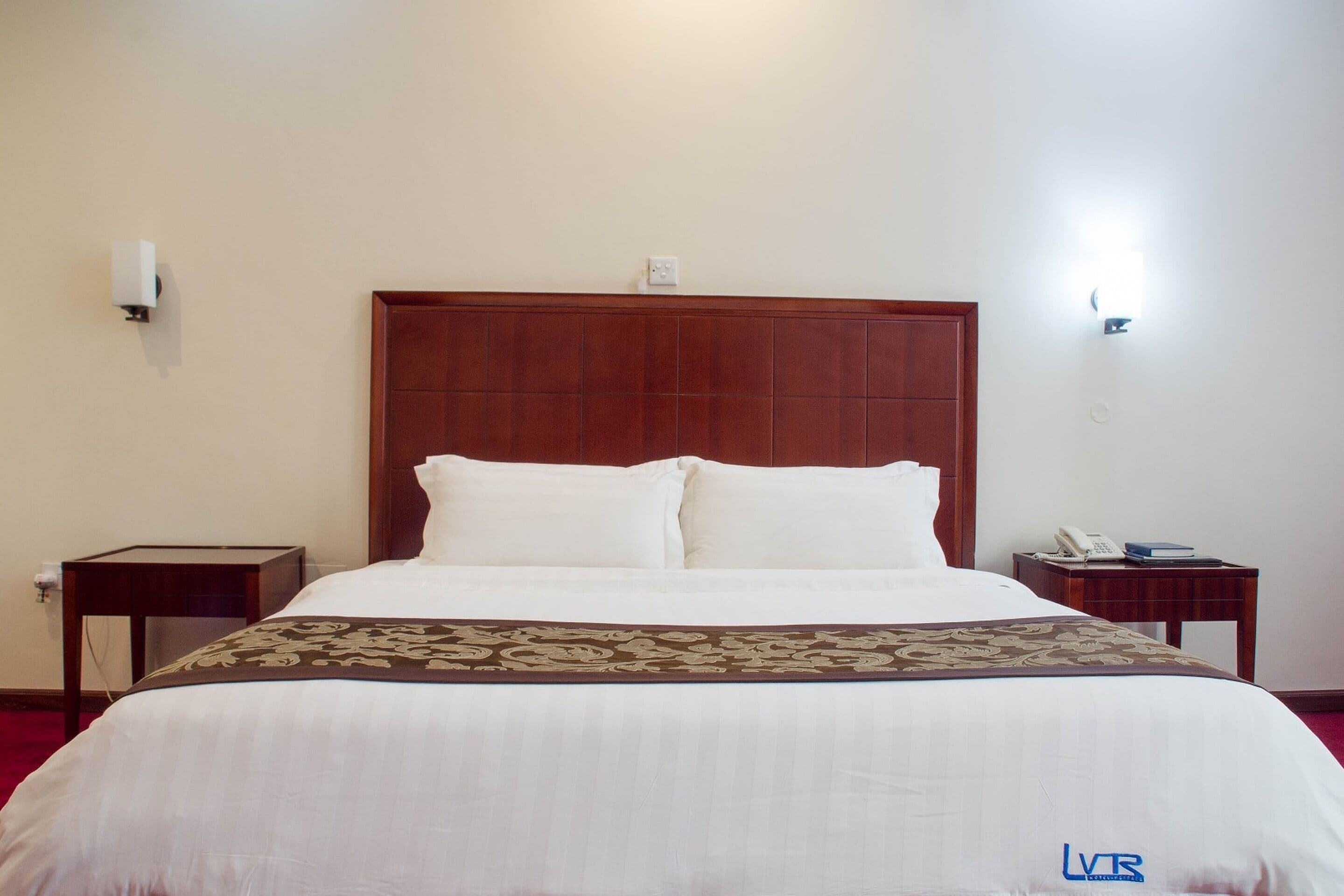 A hotel room with a large bed, a wooden headboard, two bedside tables, and a telephone on one of the tables.