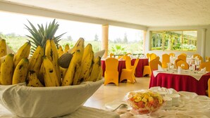 A dining area with a large bowl of bananas and a fruit bowl on a table, yellow chairs, and a view of greenery outside. - Lake View Resort Hotel (Mbarara)