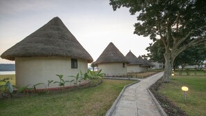 A row of thatched-roof cottages with a paved walkway and a tree in the foreground. - Victoria Forest Resort (Bugala Island)