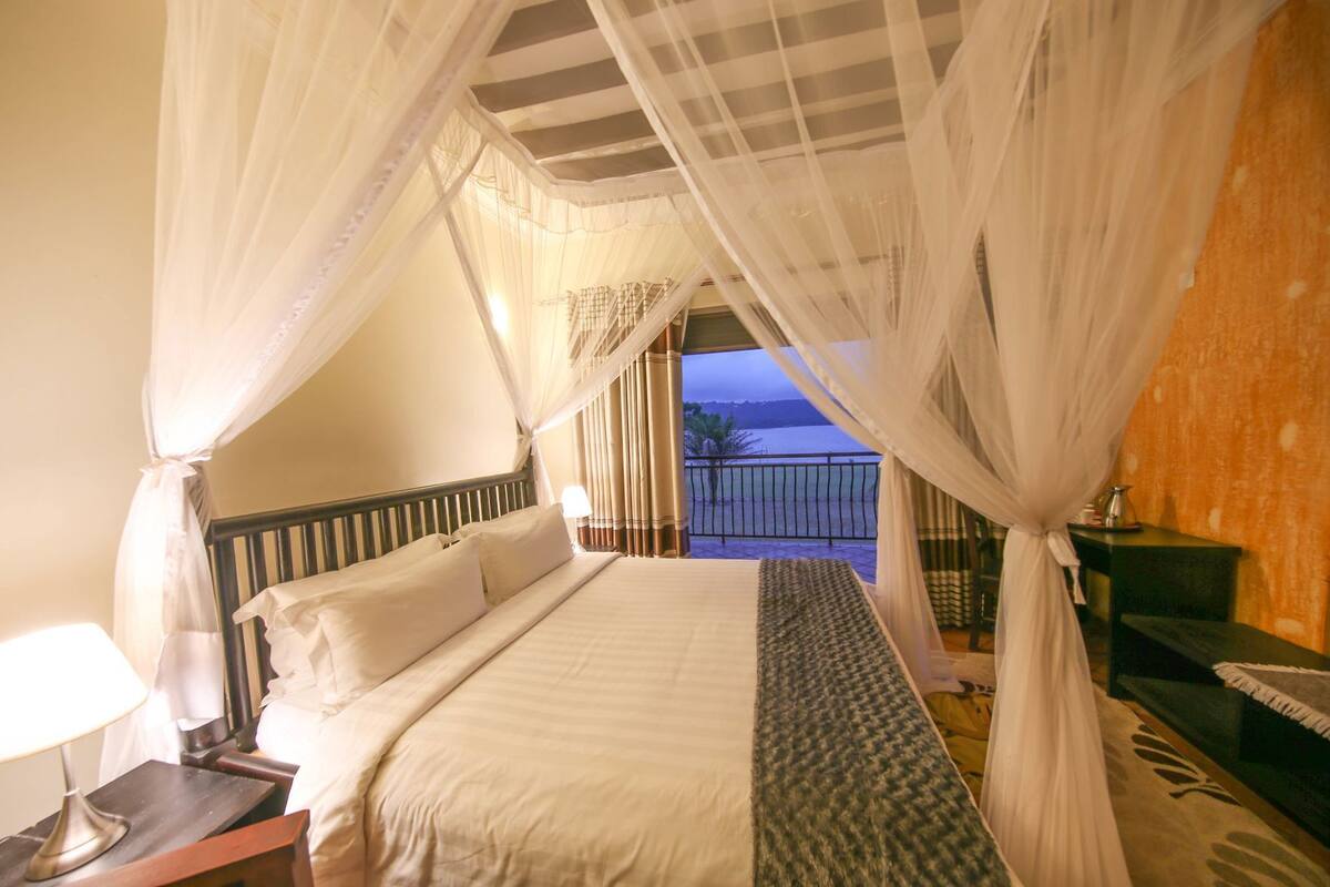 Una cama con ropa blanca, una mesita de noche con lámpara y vista al mar desde un balcón.