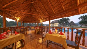 An outdoor restaurant with tables set for dining, a thatched roof, and a pool in the background. - Victoria Forest Resort (Bugala Island)