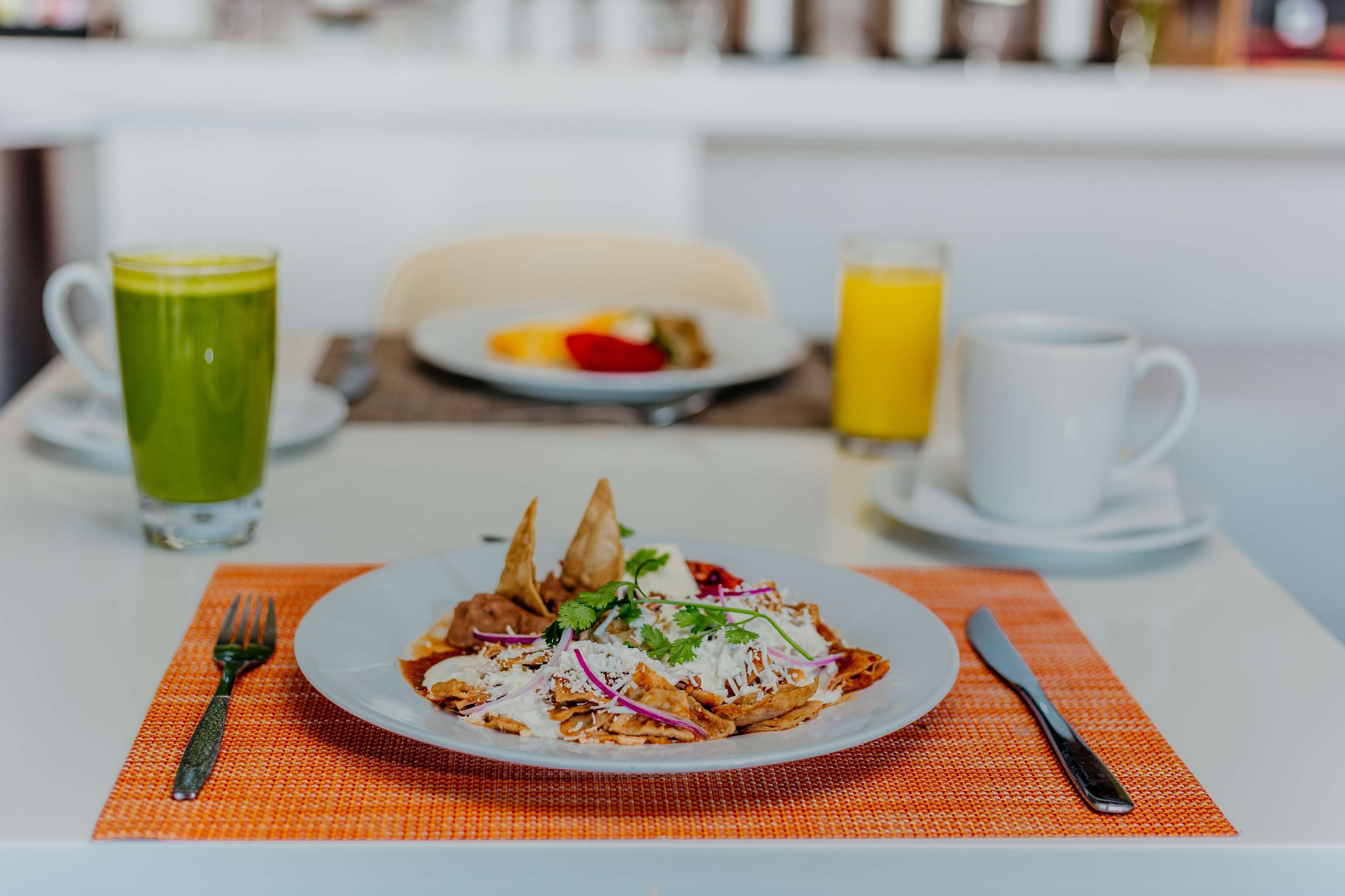 Un desayuno con una fuente de nachos, una bebida verde y un vaso de jugo de naranja sobre una mesa blanca.