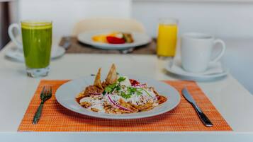 Un desayuno con una fuente de nachos, una bebida verde y un vaso de jugo de naranja sobre una mesa blanca.