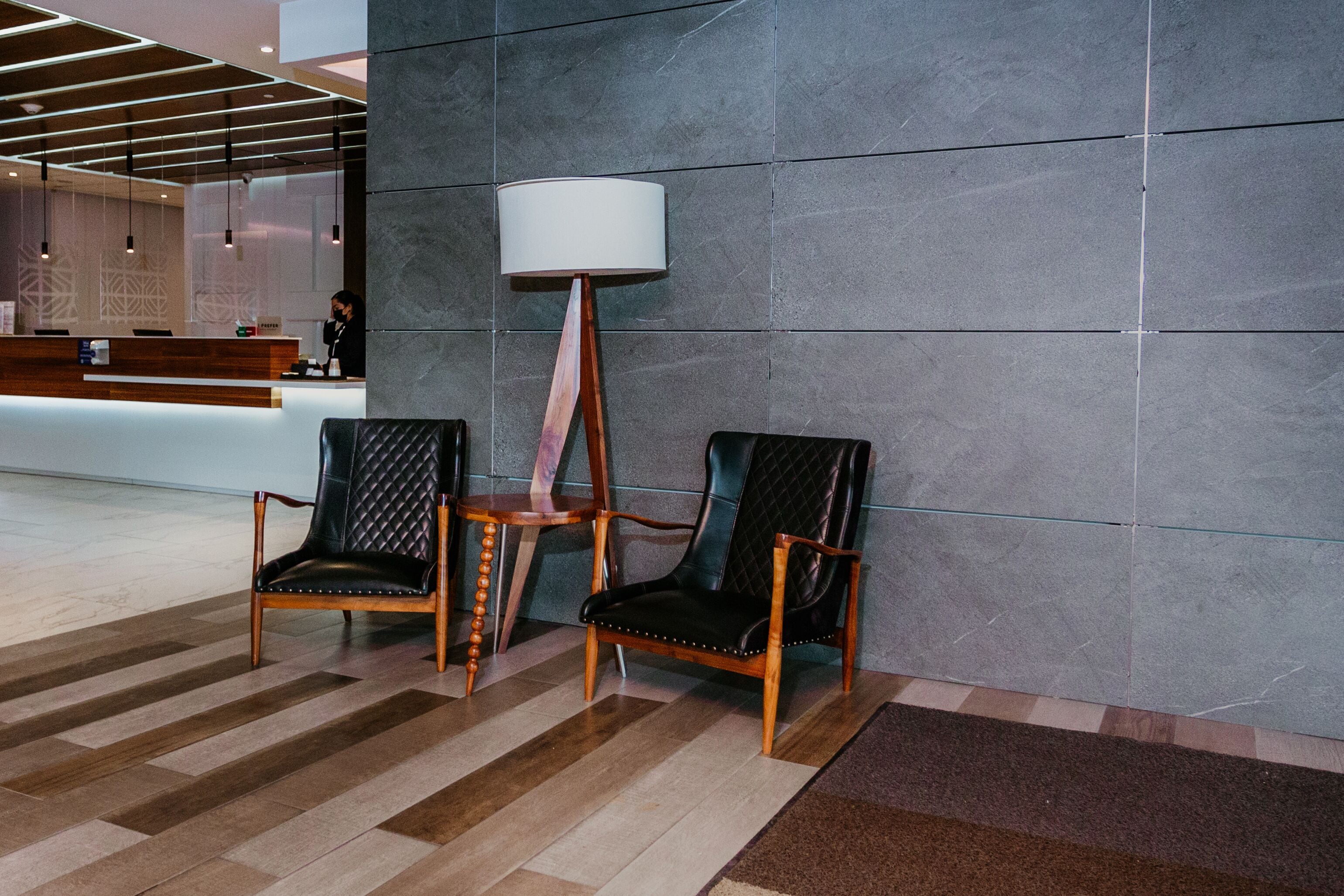 Zona de recepción con mostrador, dos sillones tapizados, lámpara de pie y alfombra estampada.