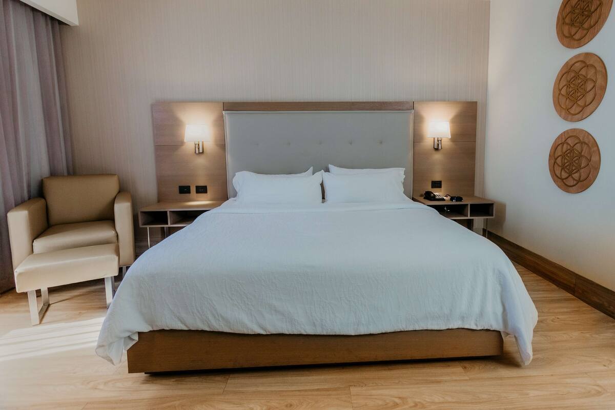 Una habitación de hotel moderna con una cama grande, dos mesitas de noche, una silla y un cuadro decorativo en la pared.